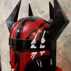 Puede incluir: Un casco negro y rojo con detalles angulares y puntiagudos. El casco tiene un acabado brillante y marcas decorativas blancas. El dise&ntilde;o recuerda a un tema de ciencia ficci&oacute;n o fantas&iacute;a.