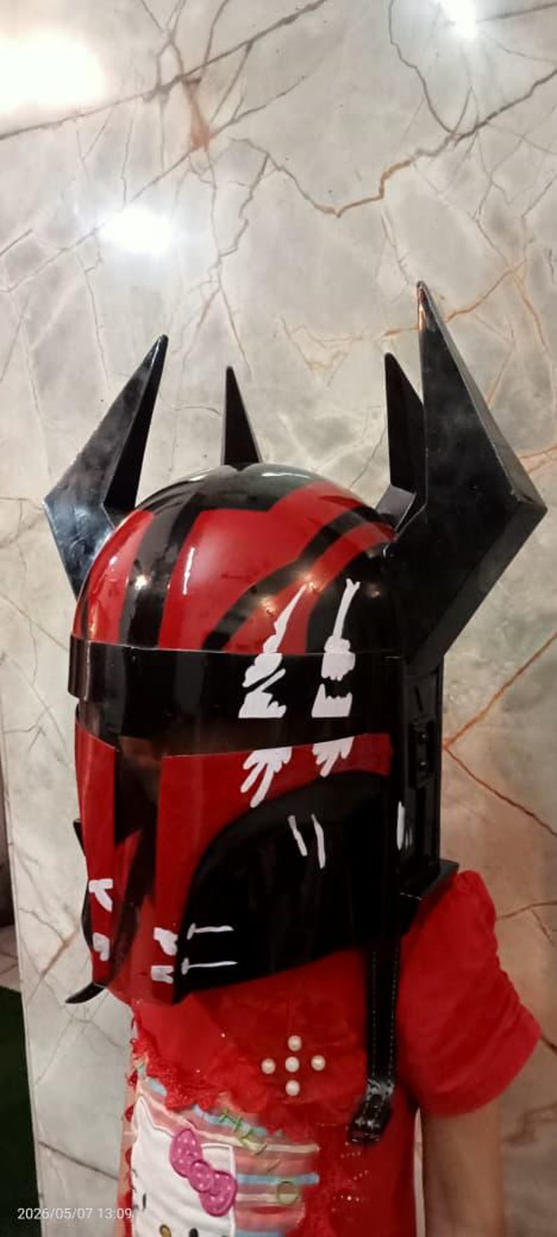 Puede incluir: Un casco negro y rojo con detalles angulares y puntiagudos. El casco tiene un acabado brillante y marcas decorativas blancas. El dise&ntilde;o recuerda a un tema de ciencia ficci&oacute;n o fantas&iacute;a.