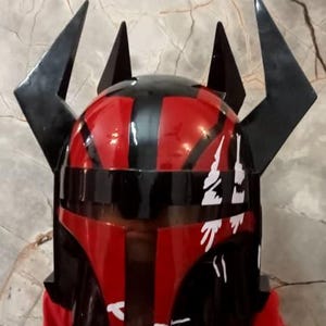 Puede incluir: Un casco rojo y negro con cuernos negros angulares. El casco tiene un acabado brillante y detalles en blanco. La imagen muestra un primer plano del casco, lo que sugiere un art&iacute;culo de disfraz o cosplay.