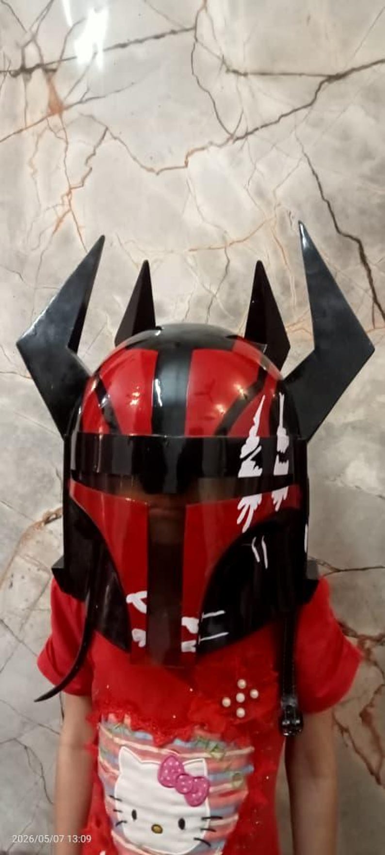 Puede incluir: Un casco rojo y negro con cuernos negros angulares. El casco tiene un acabado brillante y detalles en blanco. La imagen muestra un primer plano del casco, lo que sugiere un art&iacute;culo de disfraz o cosplay.