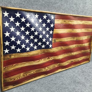 Puede incluir: Bandera americana de madera con un acabado envejecido. La bandera presenta un campo azul con estrellas blancas y rayas rojas y de madera natural. La bandera está enmarcada con un borde de madera natural. La veta de la madera es visible.