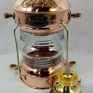 May include: A copper and brass nautical anchor lamp with a clear glass globe. The lamp has a handle and the text "ANCHOR LAMP" and "LEEDS BURTON SOUTHAMPTON" is visible. A separate brass oil burner is next to the lamp.