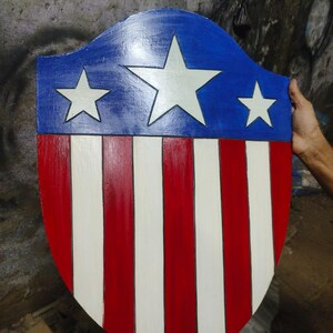 May include: A wooden shield painted in red, white, and blue. The top section is blue with three white stars. The lower section has alternating red and white vertical stripes. The shield is held by a person.