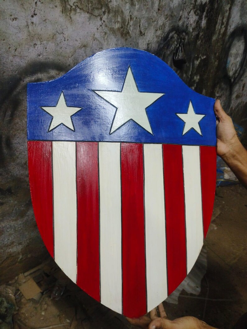 May include: A wooden shield painted in red, white, and blue. The top section is blue with three white stars. The lower section has alternating red and white vertical stripes. The shield is held by a person.