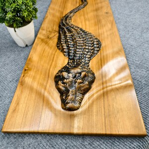 May include: A wooden wall art piece featuring a detailed relief carving of an alligator. The alligator is dark brown and textured, set against a light brown, rectangular wooden background. The artwork is a decorative piece.