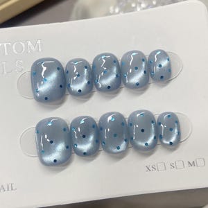 May include: A set of ten press-on nails in a light blue cat-eye finish with small blue hexagonal glitter accents. The nails are arranged in two rows on a white display card labeled CUSTOM NAILS with size options XS, S, M, and L printed at the bottom.
