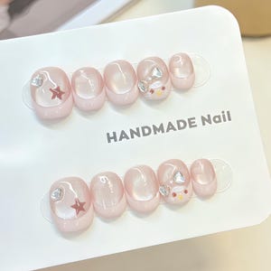 Könnte beinhalten: Ein Set aus zehn künstlichen Fingernägeln auf einer weißen Präsentationskarte mit der Aufschrift HANDMADE Nail. Die Nägel haben ein glänzendes hellrosa Finish im French-Tip-Stil. Verziert mit kleinen silbernen Sternen und Herz-Edelsteinen.