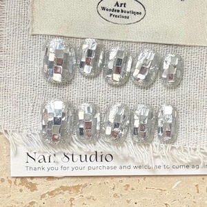 May include: Silver mosaic-style press-on nails. Each nail is covered in small, reflective squares, creating a shimmering effect. The set includes various sizes. The image also shows a tag with the text "Art Wooden boutique Precious" and a card with the text "Nar Studio".