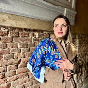 Puede incluir: Un pañuelo de seda azul con un patrón geométrico que presenta menorás, estrellas y otros símbolos. El pañuelo tiene un borde azul claro y está drapeado sobre un abrigo marrón. El fondo incluye una pared de ladrillo y detalles arquitectónicos.