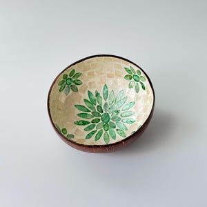 May include: A decorative bowl crafted from a coconut shell, showcasing a mosaic interior with a floral pattern in green hues. The bowl has a dark brown rim and a light-coloured mosaic background.
