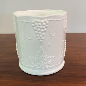 May include: A white milk glass vessel featuring a raised, embossed pattern of grape clusters and vine leaves encircling the exterior. The container has a scalloped top rim and a smooth, circular base, presenting a monochromatic design throughout.
