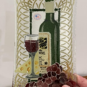 May include: Rectangular glass serving tray with a wine theme. The tray features a green wine bottle, a glass of red wine, a wedge of cheese, and a bunch of grapes. The background has a gold Celtic knot design. The words "Handmade in America" are visible.