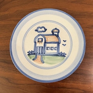 May include: A round ceramic plate with a hand-painted illustration of a house, fence, and clouds. The house has a brown roof and blue walls. The plate has a blue border and a cream-colored center. The plate is on a wooden surface.