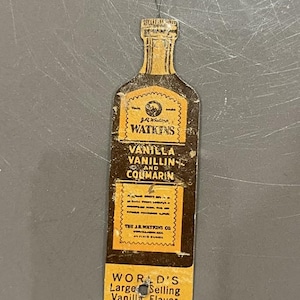 May include: Vintage Watkins Vanilla extract bottle-shaped tag. The tag is brown and gold with text that reads "Vanilla, Vanillin, and Coumarin." It also says "World's Largest Selling Vanilla Flavor."