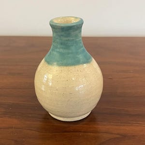 May include: A small, handcrafted ceramic vase with a rounded body and a narrow neck. The vase features a speckled, off-white base and a teal-colored top. The vase is likely used for decorative purposes.