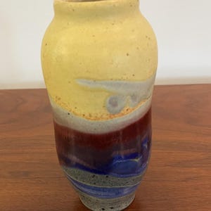 May include: A handmade ceramic vase with a cylindrical shape. The top is yellow, transitioning to white, then red, blue, and gray at the base. The vase has a speckled texture and a slightly flared rim. A decorative piece for the home.