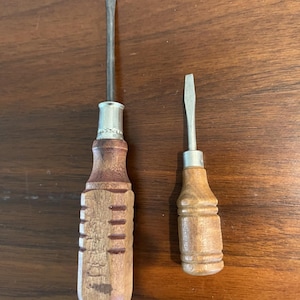 May include: Two vintage screwdrivers with wooden handles. One is larger with a ridged handle and a silver metal collar. The other is smaller with a smooth handle and a silver metal collar. Both have flathead tips.