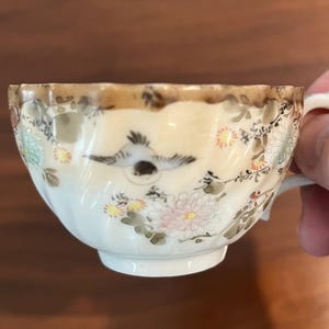 May include: A small, antique teacup with a cream-colored base and a brown rim. The cup is decorated with a flying bird and floral patterns in shades of pink, green, and yellow. The handle is small and curved.