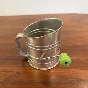 May include: A cylindrical metal flour sifter with a silver finish, featuring a side handle and a rotating crank mechanism. The crank is equipped with a bright lime green wooden knob, providing a contrast to the metallic body of the kitchen tool.