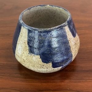 May include: A small, rounded ceramic cup featuring a speckled beige base with thick, hand-painted vertical stripes in a deep navy blue glaze. The interior of the cup is also speckled beige, and the exterior surface has a glossy, textured finish.