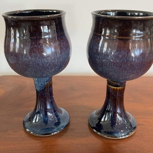 May include: Two handmade ceramic goblets with a glossy, dark blue and brown glaze. The goblets have a bulbous bowl shape and a slender stem, sitting on a wooden surface. The ceramic pieces have a unique, artistic appearance.
