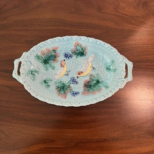 May include: An oval ceramic serving tray with a light blue glaze, featuring a raised relief design of two birds perched on branches among green and pink grape leaves and clusters of small blue grapes, with ornate scrollwork handles.