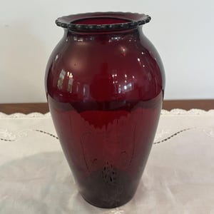 May include: A deep ruby red glass vase with a bulbous body and a narrow neck. The rim features a scalloped, crimped edge. The surface of the glass displays a faint, etched floral pattern that wraps around the lower section of the vessel.