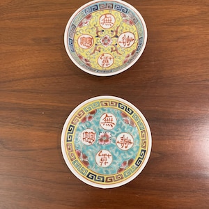 May include: Two round ceramic saucers featuring intricate patterns and Chinese calligraphy characters. The top saucer has a yellow background, while the bottom saucer has a turquoise background. Both feature a central floral motif and a geometric border.
