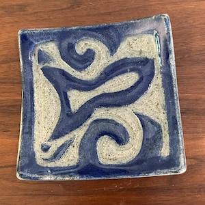 May include: Square ceramic dish with a blue and gray abstract design. The dish has a dark blue border and a textured surface. The design features swirling lines and shapes in contrasting colors, creating a unique decorative piece.