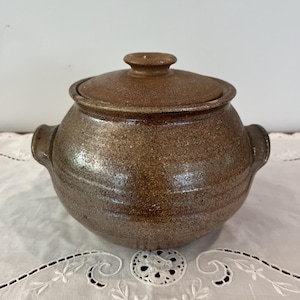 May include: A round, brown ceramic pot with a speckled, textured finish. The pot features two side handles, a wide body, and a matching lid with a central knob. The surface has subtle horizontal ridges circling the exterior of the vessel.