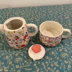 May include: A ceramic tea set with a floral pattern, comprising a teapot, a teacup, and a lid. The teapot and teacup feature a white base with red and pink floral designs. The lid has a red flower-shaped handle. The set is on a blue floral tablecloth.