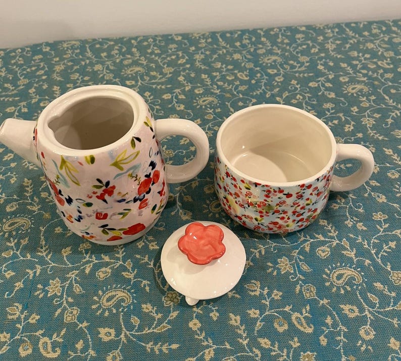 May include: A ceramic tea set with a floral pattern, comprising a teapot, a teacup, and a lid. The teapot and teacup feature a white base with red and pink floral designs. The lid has a red flower-shaped handle. The set is on a blue floral tablecloth.
