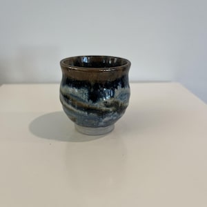 May include: A small, handmade ceramic cup with a unique, textured design. The cup features a dark brown rim, transitioning to shades of blue and white, and a light gray base. The cup is likely used for drinking.