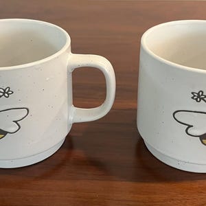 May include: Two white ceramic mugs with a speckled finish. Each mug features a whimsical bee design with black outlines, yellow and black stripes, and small flower accents. The mugs have a simple handle and a slightly flared rim.