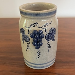 May include: A beige ceramic jar with a blue grape design. The jar has a cylindrical shape with a slightly flared rim and a blue band around the top. The grape design includes a cluster of grapes and leaves.
