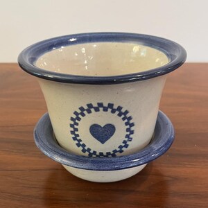 May include: A ceramic planter with a matching saucer. The planter is off-white with a blue rim and a blue heart design in the center. The saucer has a blue rim. The planter and saucer are sitting on a wooden surface.