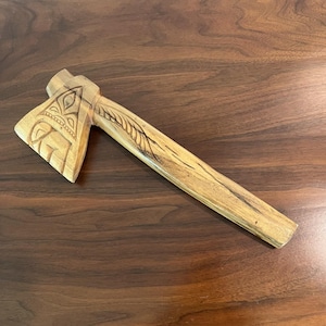 May include: A hand-carved wooden axe with intricate designs on the head and handle. The axe head features geometric patterns, while the handle has a leaf-like carving. The wood has a natural, light brown color.