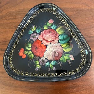 May include: A black, triangular serving tray with a floral design. The tray features a gold trim and a hand-painted bouquet of red, pink, and white roses with green leaves. The tray is on a wooden surface.