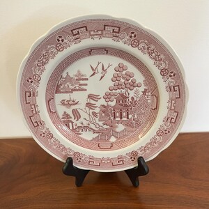 May include: A round ceramic plate featuring a maroon and white transferware pattern. The design depicts a landscape with a pagoda, a bridge, a boat, and two birds in flight. The rim is decorated with a geometric border pattern.