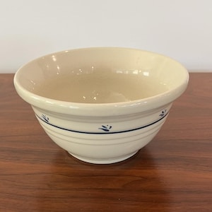 May include: Cream-colored ceramic mixing bowl with a blue floral design. The bowl has a wide rim and a rounded shape. A blue stripe and floral accents encircle the bowl. The bowl is sitting on a wooden surface.