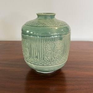 May include: A celadon green ceramic vase with a round body and a narrow neck. The vase features embossed floral and vertical line patterns. The base and the area below the neck have a wave-like pattern.