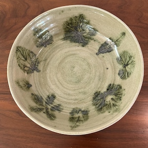 May include: A handmade ceramic bowl with a light green glaze. The bowl features a leaf design in shades of green and gray. The leaves are arranged around the bowl's interior. The bowl is round and has a slightly raised rim.