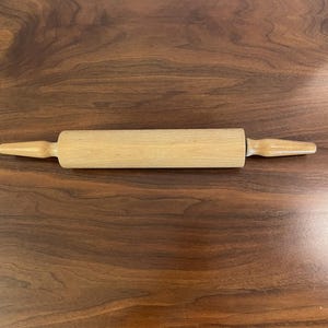 May include: A light brown wooden rolling pin featuring a smooth cylindrical barrel and two tapered handles on either side. The kitchen tool is crafted from a single piece of light-colored wood with a visible natural grain pattern throughout.