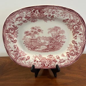 May include: An oval serving platter with a detailed floral and landscape design in burgundy and white. The rim features a dotted border and floral patterns. The center showcases a scenic landscape with a bridge and buildings. The platter is displayed on a black stand.