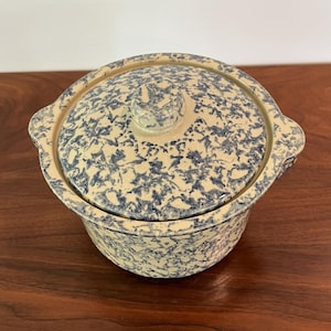 May include: A ceramic casserole dish with a lid. The dish and lid are cream-colored with a blue floral pattern. The dish has two handles and a knob on the lid. The dish is sitting on a dark wooden surface.