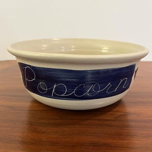May include: Cream-colored ceramic popcorn bowl with a dark blue band. The word "Popcorn" is written in cursive across the blue band. The bowl has a rounded shape and is sitting on a wooden surface.