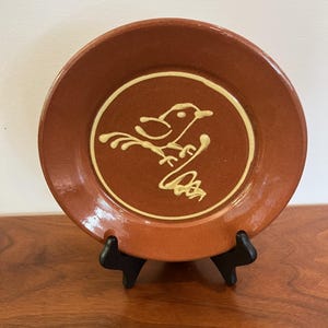 May include: A round terracotta-colored ceramic plate featuring a raised, light-colored line drawing of a bird perched on a branch in the center. The plate has a smooth, glossy finish and is displayed on a black plastic stand for upright viewing.