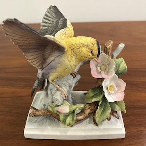May include: A ceramic figurine of a yellow bird with black and gray wings perched on a branch with pink and white flowers. The bird is in mid-flight, with its wings spread. The base is white and rectangular.
