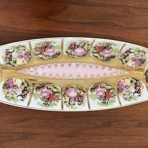 May include: Oval serving dish with a white base, featuring a pink center and gold trim. The dish is decorated with scenes of people in period clothing. The rim has a gold border, and the dish is set against a wooden surface.
