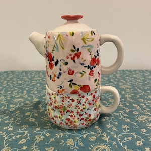 May include: A ceramic teapot and teacup set, stacked together. The teapot is pale pink with a floral pattern of red, blue, and yellow flowers. The teacup has a similar floral design on a light blue background. Both have white handles.